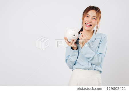 Excited happy Asian woman holding piggy bank while pointing finger to piggy savings plan focused on money goals and believing in future banking stability. isolated studio white background 129802421