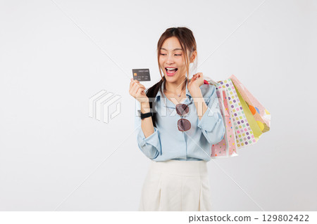 Happy Asian woman showing shop finance credit card, holding colorful shopping bags fresh from the store and presents her credit card with excitement after payment, headshot isolated white background 129802422