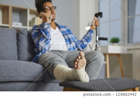 Man talking on phone while sitting on couch, with his injured foot resting on stool 129802442