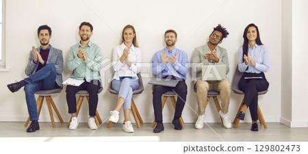 Group of professional business people sitting on chairs in row looking at camera and applauding. 129802473
