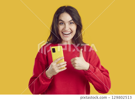 Happy woman sincerely rejoices for good news shows thumb up with smartphone on yellow background. 129802513