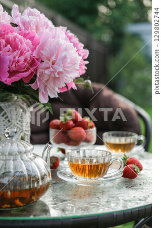 Summer still life with vase of pink peonies, glass teapot and two cups of tea on garden table. Suitable for themes of summer, cottagecore, relaxation and healthy eating 129802744