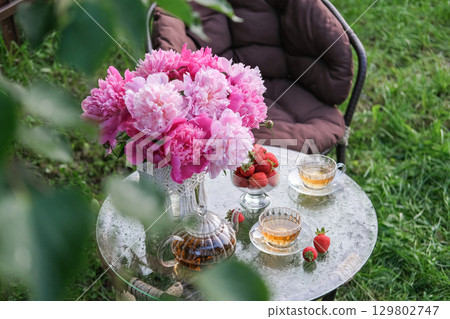 Charming summer garden scene featuring glass table with vase of pink peonies, glass teapot, two cups of tea and bowl of fresh strawberries. Suitable for themes of summer, tea time, relaxation 129802747