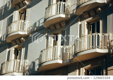 A building with elegant balconies and architectural details in a light. Facade with balconies, ornate railings, and open shutters on the windows. Classic European (Spanish) architecture. 129803304