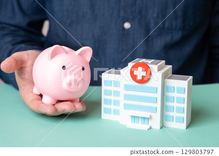 A man holds a piggy bank near a hospital building. Medical insurance. Patient care funding. 129803797