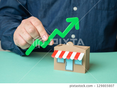 A man holds a green arrow pointing up over a store. Growing store profits, expanding A man holds a green arrow pointing up over a store. Growing store profits, expanding 129803807