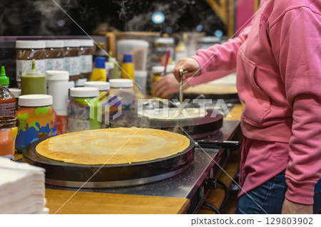 Street food, pancakes. Cook prepares hot pancakes on a street food stall surrounded by toppings and condiments. Concept of pancake making, winter street food, cozy festive flavors 129803902