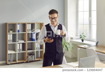 Man standing with a cup coffee at office and reading messages on his mobile phone. 129804047