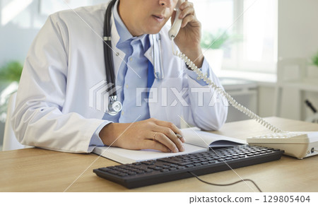 Medical professional doctor, male nurse at office desk, talking on phone, consulting notebook Medical professional doctor, male nurse at office desk, talking on phone, consulting notebook 129805404