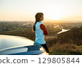 Young woman standing near her car enjoying warm sunset view. Girl traveler leaning on vehicle hood looking at evening horizon. 129805844