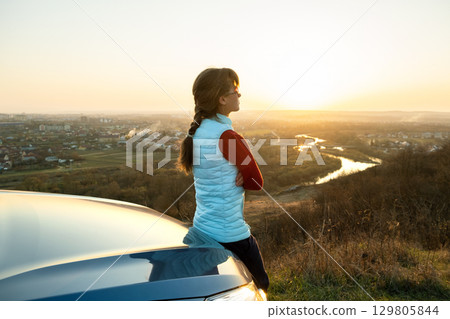 Young woman standing near her car enjoying warm sunset view. Girl traveler leaning on vehicle hood looking at evening horizon. 129805844