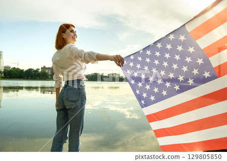 Happy young womanl with USA national flag on her shoulders with high city buildings in background celebrating United States independence day. Happy young womanl with USA national flag on her shoulders with high city buildings in background celebrating United States independence day. 129805850