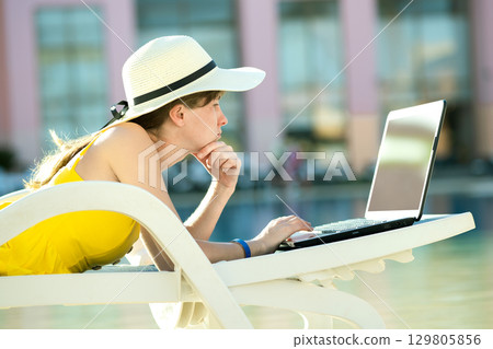 Young woman in yellow dress is laying on beach chair working on computer laptop connected to wireless internet typing text on keys in summer resort. Doing studies while travelling concept. 129805856