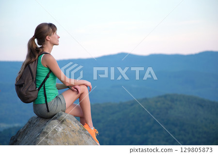 Tired hiker girl relaxing on rocky mountain top enjoying summer nature during travelling on wilderness trail. Lonely woman traveler traversing high hilltop route. Healthy lifestyle concept Tired hiker girl relaxing on rocky mountain top enjoying summer nature during travelling on wilderness trail. Lonely woman traveler traversing high hilltop route. Healthy lifestyle concept 129805873