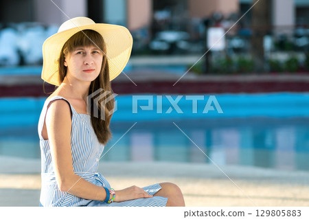 Young woman wearing light summer dress and yellow straw hat sitting outside near hotel swimming pool on summer sunny day. 129805883