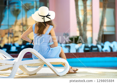 Young woman wearing light summer dress and yellow straw hat sitting outside near hotel swimming pool on summer sunny day. 129805884