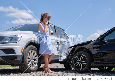 Stressed driver talking on sellphone on roadside near her smashed vehicle calling for emergency service help after car accident. Road safety and insurance concept 129805891