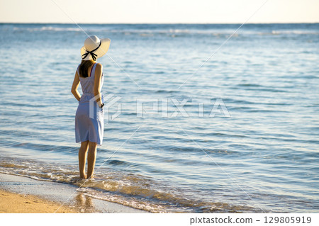 Young woman in straw hat and a dress standing alone on empty sand beach at sea shore. Lonely tourist girl looking at horizon over calm ocean surface on vacation trip. 129805919