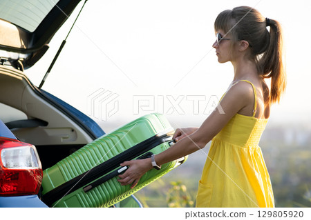 Young woman in summer dress putting green suitcase inside her car trunk. Travel and vacations concept. 129805920