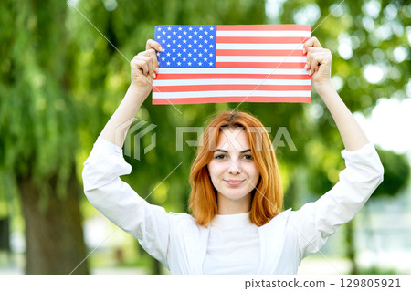 Happy red haired girl posing with USA national flag up over her head standing outdoors in summer park. Positive young woman celebrating United States independence day. 129805921