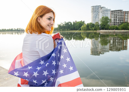 Happy smiling young womanl with USA national flag on her shoulders with high city buildings in background celebrating United States independence day. 129805931