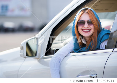 Young fashionable smiling woman driver looking out the window behind car steering wheel. 129805954
