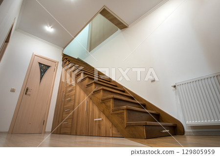 Modern architecture interior with luxury hallway with glossy wooden stairs in multi-storey house. Custom built pullout cabinets on glides in slots under stairs. Use of space for storage. 129805959