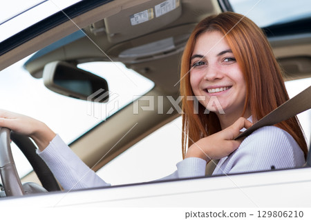 Wide angle view of young redhead woman driver fastened by seatbelt driving a car smiling happily. 129806210