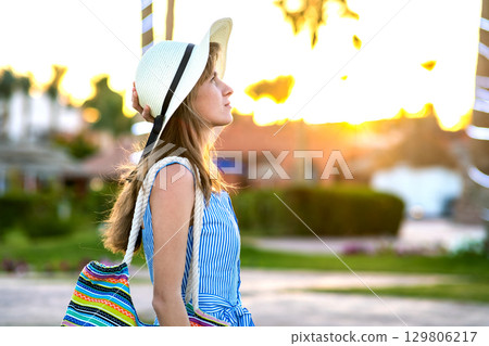 Young woman wearing light blue summer dress and yellow straw hat holding fashionable shoulder bag standing outside enjoying warm weather in summer park at sunset. 129806217