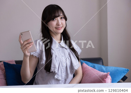客廳裡，一位面帶微笑的日本女性手裡拿著智慧型手機 129810404