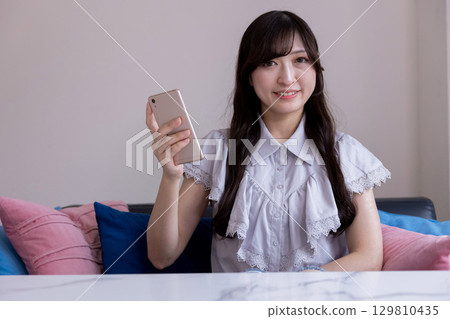 客廳裡，一位面帶微笑的日本女性手裡拿著智慧型手機 129810435