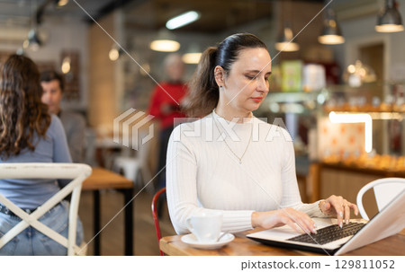 Woman surfing Internet on notebook while drinking tea or coffee in cafeteria Woman surfing Internet on notebook while drinking tea or coffee in cafeteria 129811052