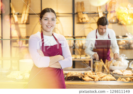 Smiling young waitress, bakery saleswoman in apron looking at the camera during working day in cafe Smiling young waitress, bakery saleswoman in apron looking at the camera during working day in cafe 129811149