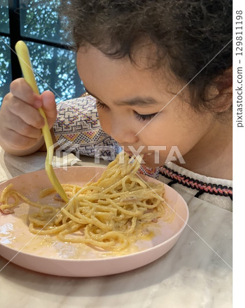 Close up view of little girl eating delicious spaghetti carbonara with a fork in a restaurant. 129811198