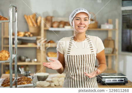 Smiling young female baker inviting to warm bakery Smiling young female baker inviting to warm bakery 129811268