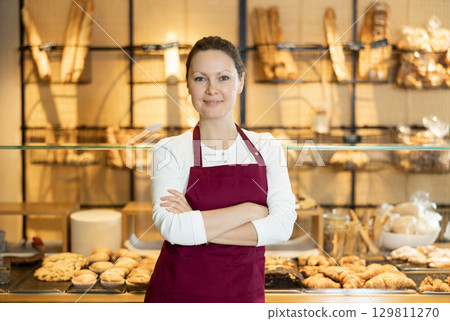 Positive middle-aged female business owner in apron standing in front of pastry stand in bakery shop. Concept of small local business Positive middle-aged female business owner in apron standing in front of pastry stand in bakery shop. Concept of small local business 129811270
