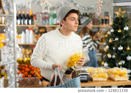 Male shopper choosing food and drinks in supermarket for christmas celebration. In background, female salesperson decorates Christmas tree with toys 129811336
