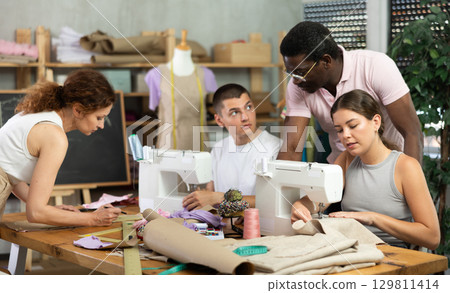 During lesson, African male teacher helps student sew, while others sew, work draw dress pattern During lesson, African male teacher helps student sew, while others sew, work draw dress pattern 129811414