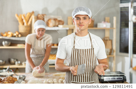 Young baker standing with dough scraper in small bakery 129811491