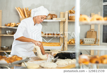 Young woman works as baker, mold baguette from dough, creates long loafs from dough. Young woman works as baker, mold baguette from dough, creates long loafs from dough. 129811539