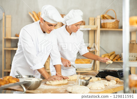 Girl and guy bakers cut small pieces of dough from large lump and put them on scales. Girl and guy bakers cut small pieces of dough from large lump and put them on scales. 129811591