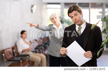 Upset man standing with papers in office scolded by angry female boss Upset man standing with papers in office scolded by angry female boss 129811645