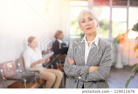 Smiling secretary senior woman stands at entrance to office reception and expects visitors 129811676