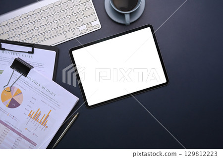 Office workspace featuring digital tablet with blank screen and financial reports on dark desk background 129812223