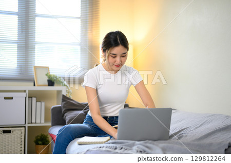 Young Asian woman working from home on a laptop while sitting on sofa Young Asian woman working from home on a laptop while sitting on sofa 129812264