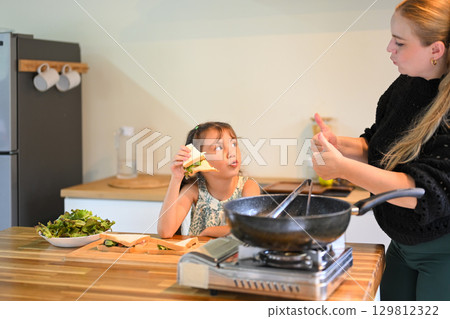 Little girl eating a homemade sandwich while her mother gives a thumbs up in the kitchen Little girl eating a homemade sandwich while her mother gives a thumbs up in the kitchen 129812322