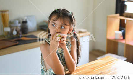 Cute little girl eating a fresh green apple in the kitchen, enjoying a healthy snack 129812336