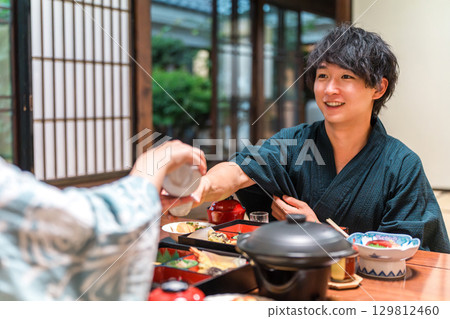 一位身穿浴衣的年輕亞洲男子在日式旅館享用晚餐時,正在享用飲料 一位身穿浴衣的年輕亞洲男子在日式旅館享用晚餐時,正在享用飲料 129812460