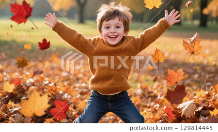 Happy little boy playing in fallen autumn leaves, arms raised with joyful expression on a sunny fall day in the park. 129814445