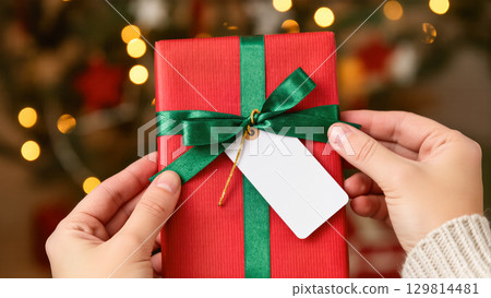 Christmas gift in red wrapping with green ribbon and tag held by hands against festive background with warm lights Christmas gift in red wrapping with green ribbon and tag held by hands against festive background with warm lights 129814481